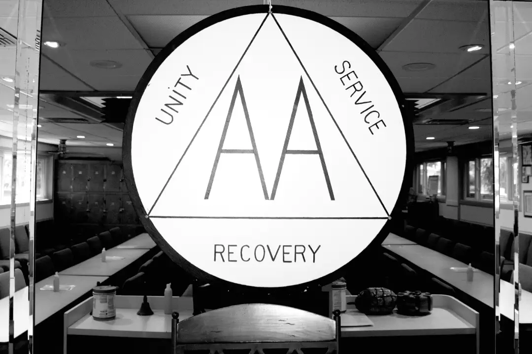 Alcoholics Anonymous symbol featuring _Unity, Service, Recovery_ in a meeting room with chairs, tables, and an American flag visible Alcoholics Anonymous symbol featuring _Unity, Service, Recovery_ in a meeting room with chairs, tables, and an American flag visible