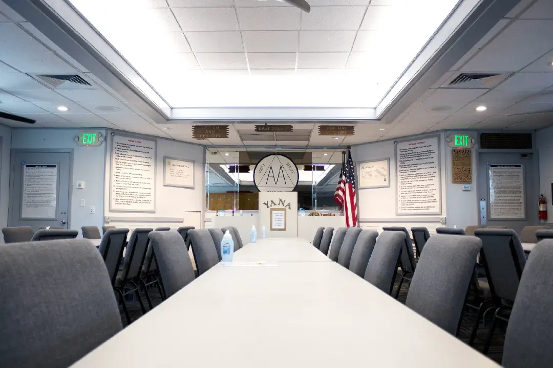 Interior of a meeting room at Y.A.N.A. with long conference tables, gray chairs, framed posters on the walls, AA signage, and an American flag near the podium Interior of a meeting room at Y.A.N.A. with long conference tables, gray chairs, framed posters on the walls, AA signage, and an American flag near the podium